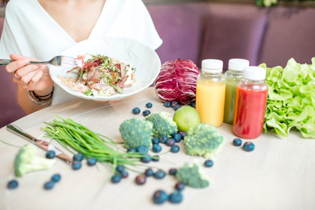 Una mujer con una ensalada en un plato y sobre la mesa arándanos, brócoli, coliflor, lechuga y tres botes con zumos de color amarillo, verde y rojo.